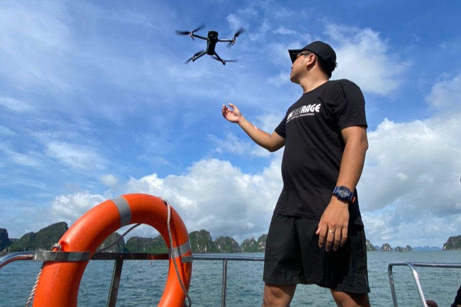 Man on a boat launches drone over sea, sunny sky. Wears black, text on shirt. Orange lifebuoy in view. Relaxed mood.