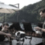 Two men setting up a drone on a patio table, surrounded by lounge chairs and umbrellas. Scenic mountain and water view in the background.