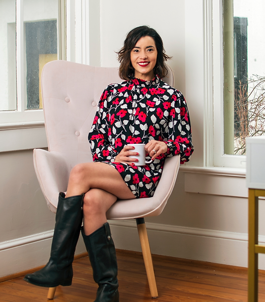 Yoli Dick, neuroscience marketing consultant and personal brand strategist, sits confidently in a modern armchair holding a white coffee mug. She wears a floral-patterned dress with black boots, smiling warmly in a bright, stylish office space—reflecting her creative, bold, and approachable brand.