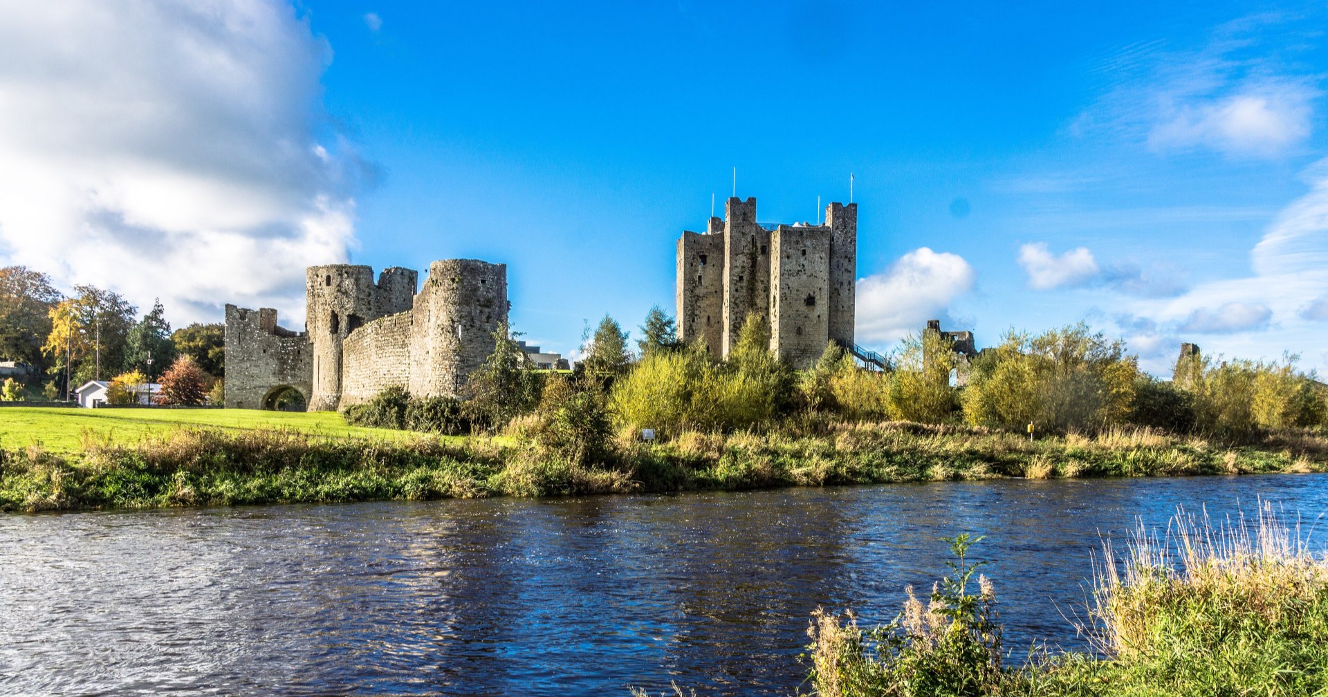 Trim Castle seen across the River Boyne on a bright autumn day: the tall square keep and the curtain wall stand above the far bank, with trees in autumn colour along the water and a blue sky with white clouds.