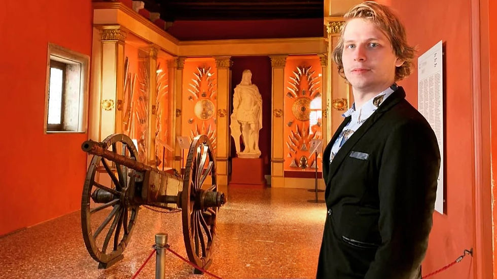 Aleksander Traks, wearing a black blazer, stands in a historical museum with a cannon and medieval weapons behind him. The warm orange lighting and classical statues evoke themes of history, learning, and mastery.