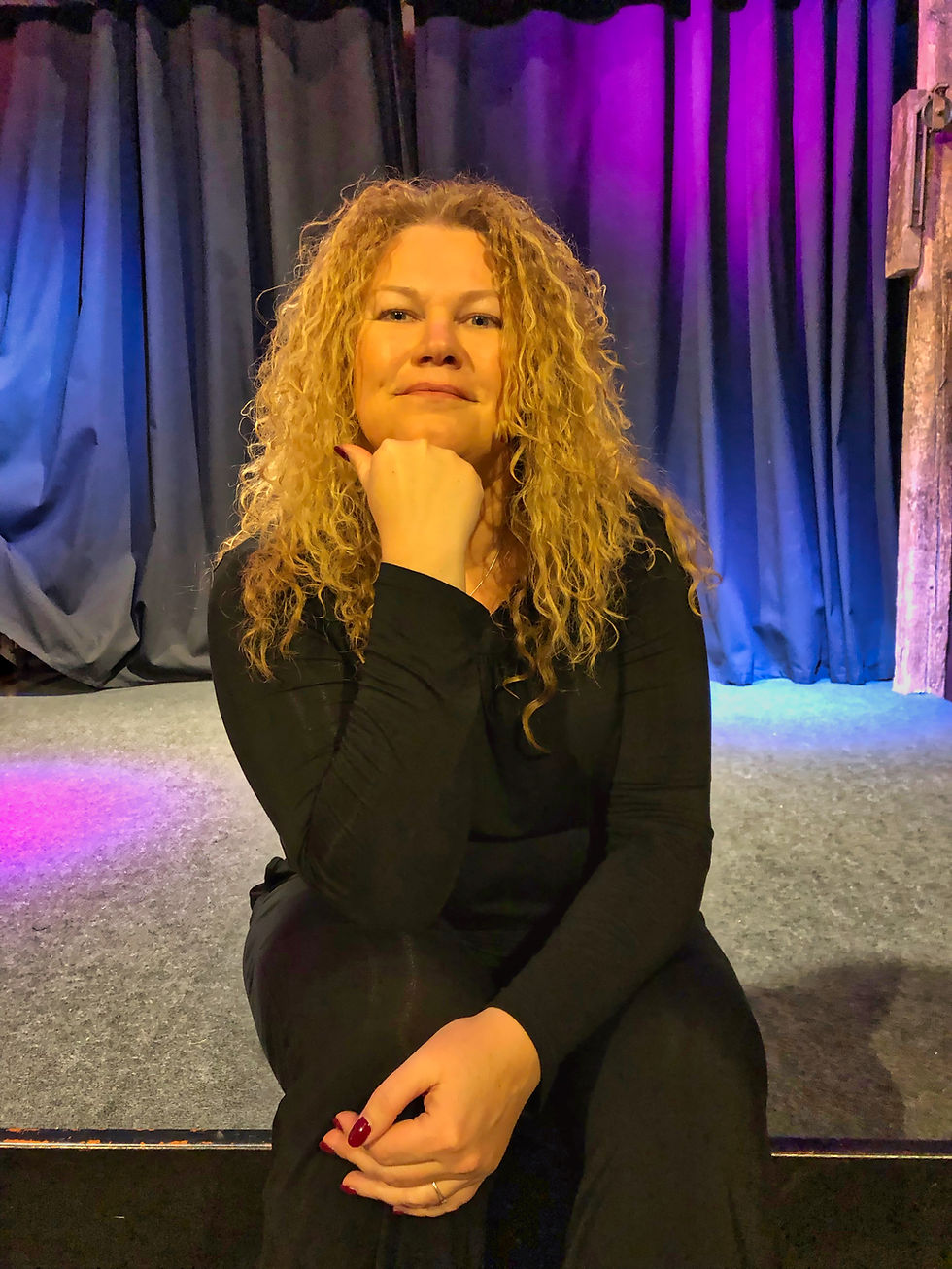 Yvonne Dibowski-Zanera mit lockigen blonden Haaren sitzt auf einer Bühne vor violett-blauen Vorhängen und blickt nachdenklich in die Kamera.