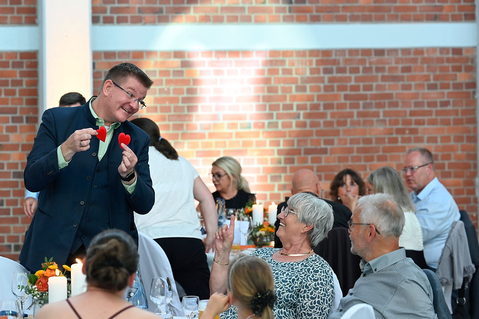 Marc Dibowski im dunkelblauen Anzug zeigt zwei rote Herzen bei einer Feier; Gäste lachen an Tischen vor Backsteinwand.