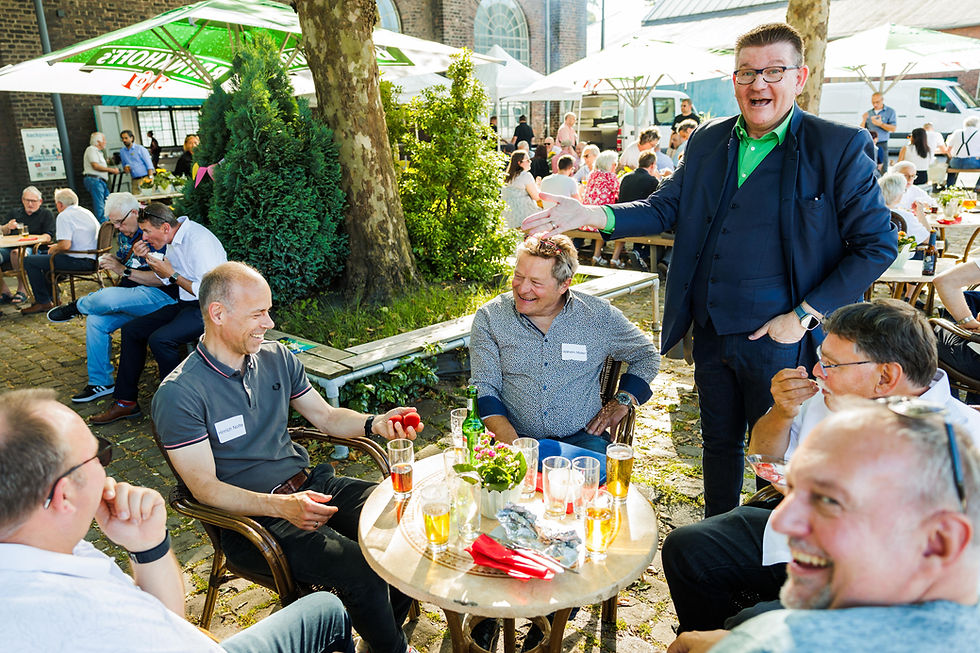 Bei einem sonnigen Gartenfest plaudern Männer an Tischen; ein Marc Dibowski im Anzug begrüßt lachend die Runde, mit Zauberkunst und Mentalmagie zum Sommerfest.