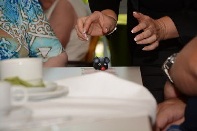 Hände greifen nach kleinem schwarzem Spielzeug auf weiß gedecktem Tisch, im Hintergrund sitzen Personen. Zaubern lernen.