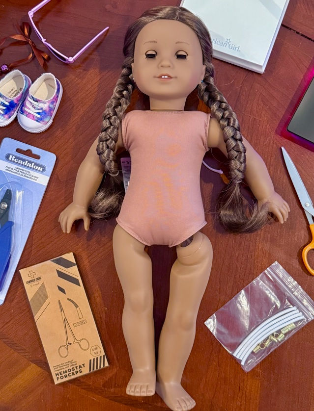 American Girl Kanani doll laying on the table with restringing tools surrounding her. One leg is dangling from its socket.
