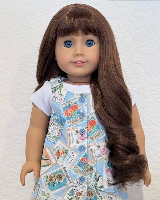 Just Like You #7 with a long brunette wig. It has bangs and the ends are curly. It's very thick. The doll is wearing a blue jumper dress with seed packets on it and a white t-shirt underneath.