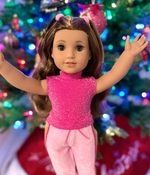 Lila doll posing with her arms raised. She wears her meet outfit (magenta top with baby pink sporty pants) and behind her is a lit Christmas tree.