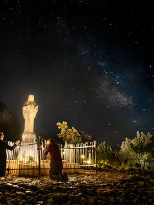 Estátua de mármore branco de Nossa Senhora de Medjugorje iluminada à noite na Colina das Aparições (Monte Podbrdo) sob um céu estrelado.
