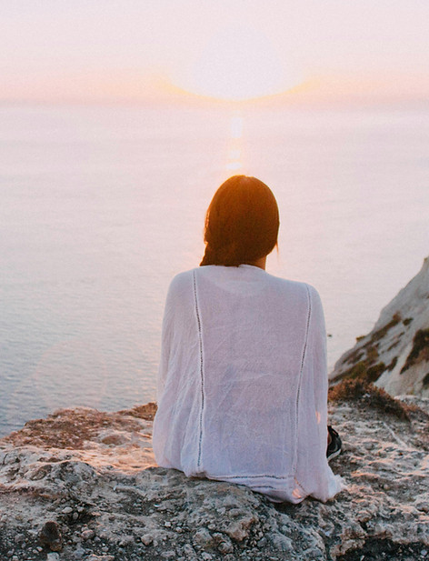 Woman in a white shirt sits on a rocky cliff, overlooking a calm sea at sunset. The mood is peaceful and contemplative.