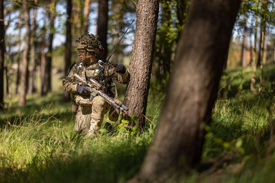 Soldier in combat gear holding a weapon in a forest UK setting.