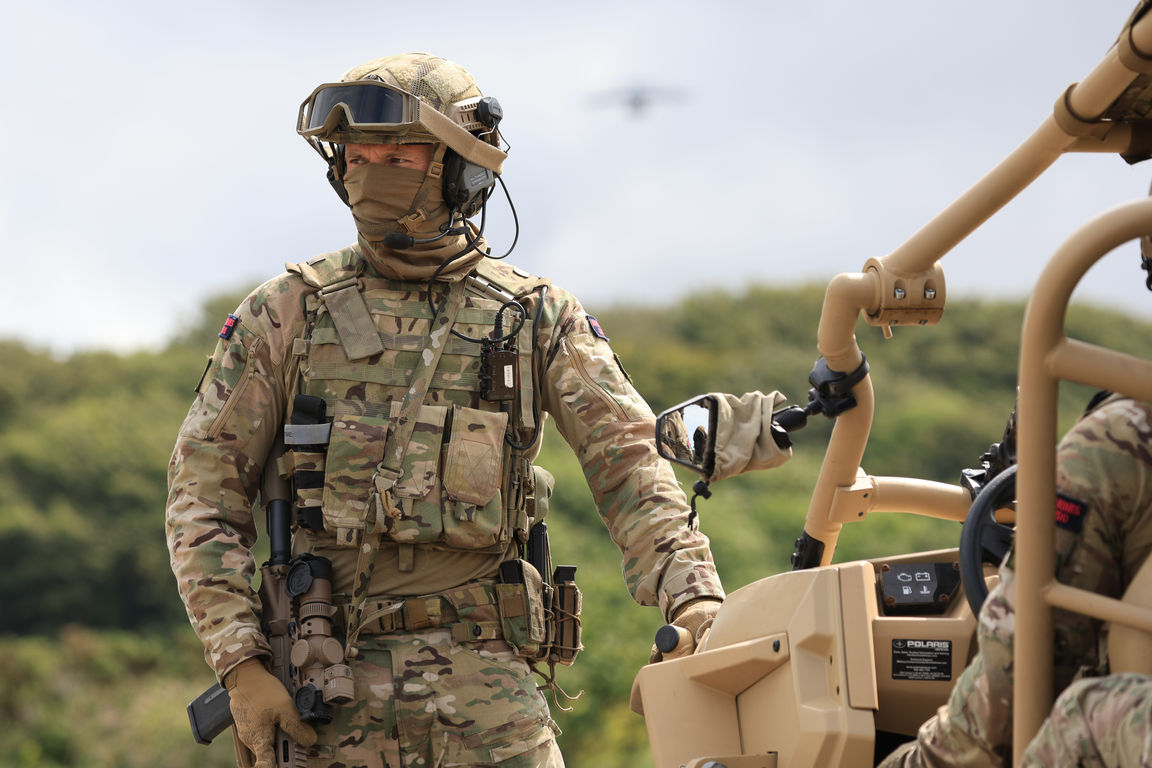 Soldier in camouflage uniform and helmet stands near a vehicle, UK background