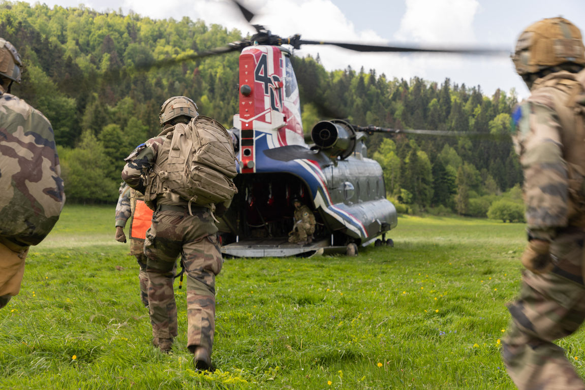 Soldiers running towards a Chinook helicopter with UK markings near trees in field.