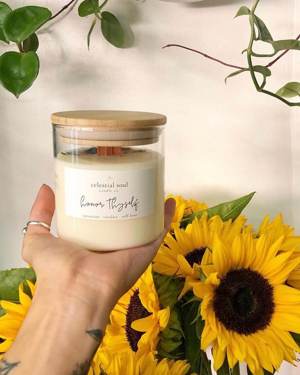 Hand holding a candle labeled "celestial soul" amid sunflowers and green vines. Bright, natural setting with a calm, uplifting mood.