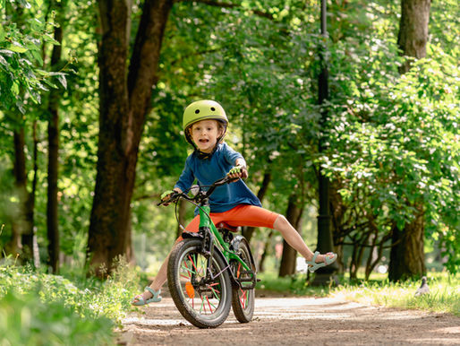 Child in a helmet rides a bike on a forest path, smiling with one leg outstretched. Lush green trees surround the scene.