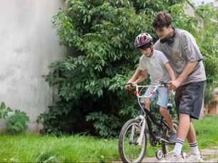 Boy learning to ride a bike with older person's help. Both focused, wearing casual clothes. Green plants and grass in the background.