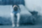 Person sitting on a bed, head in hands, visible through a rain-streaked window. The scene is moody with blue and gray tones.