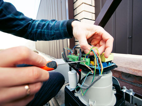 Hands using a screwdriver to repair wiring on an outdoor electrical device beside a building, with multicolored cables and a focused mood