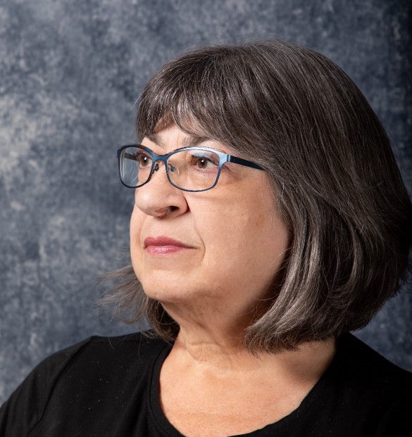 Studio portrait of Robyn Rennie, wearing blue-framed glasses and a black top, with shoulder-length grey and dark hair, looking to the side against a grey backdrop.