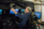 Man operating a flight simulator, adjusting controls. Blue screen showing runway scene. Monitors display data. Focused atmosphere.