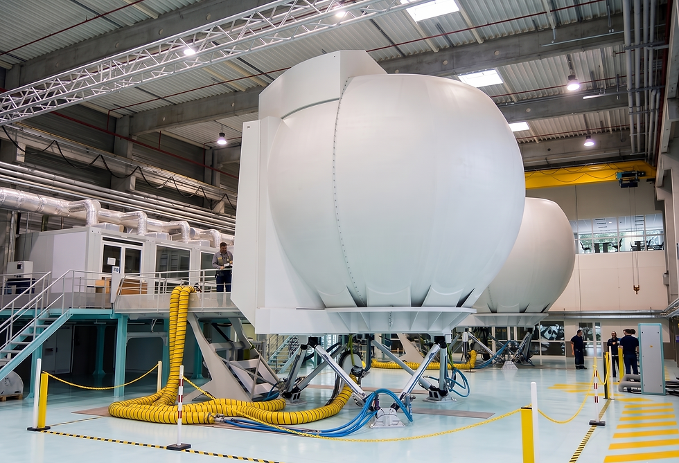 Large white flight simulators in an industrial building. Yellow hoses attached. Technicians work nearby. Overhead lights illuminate the scene.