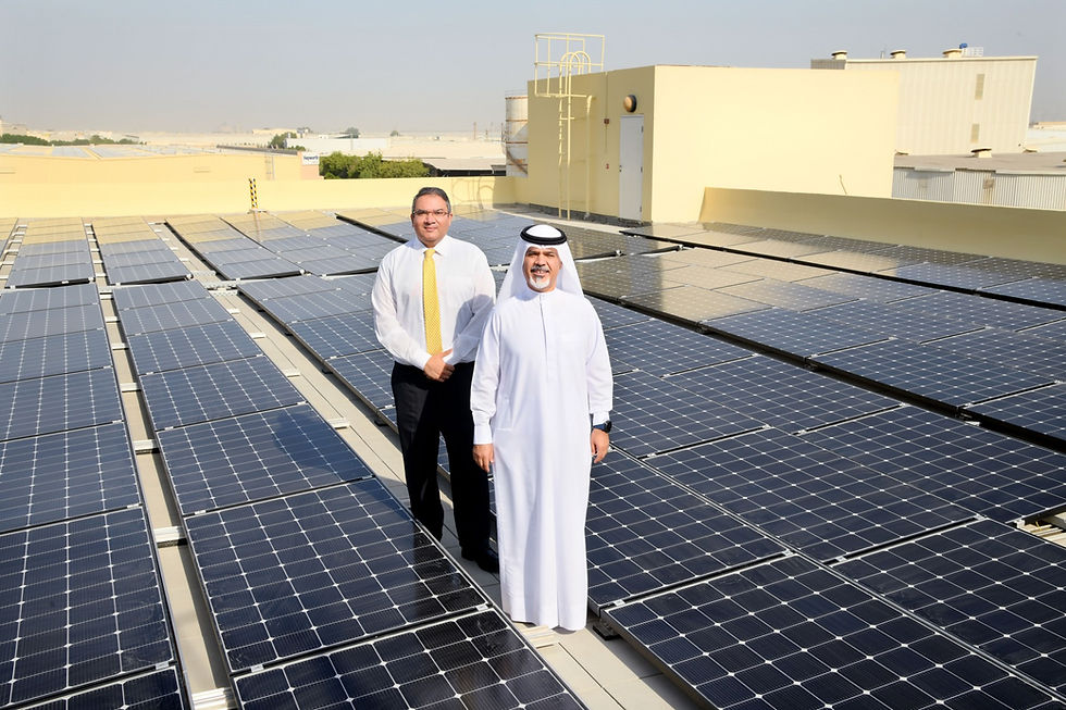 Two men stand on a rooftop solar panel array, one in a white kandura and one in a shirt and tie, with industrial buildings behind.