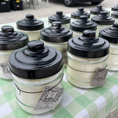 Jars of candles with black lids, displayed on a checkered tablecloth at a market.