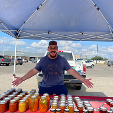 A happy vendor at a market stand displaying jars of homemade preserves under a blue canopy.
