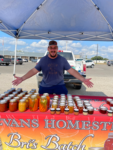 A happy vendor at a market stand displaying jars of homemade preserves under a blue canopy.