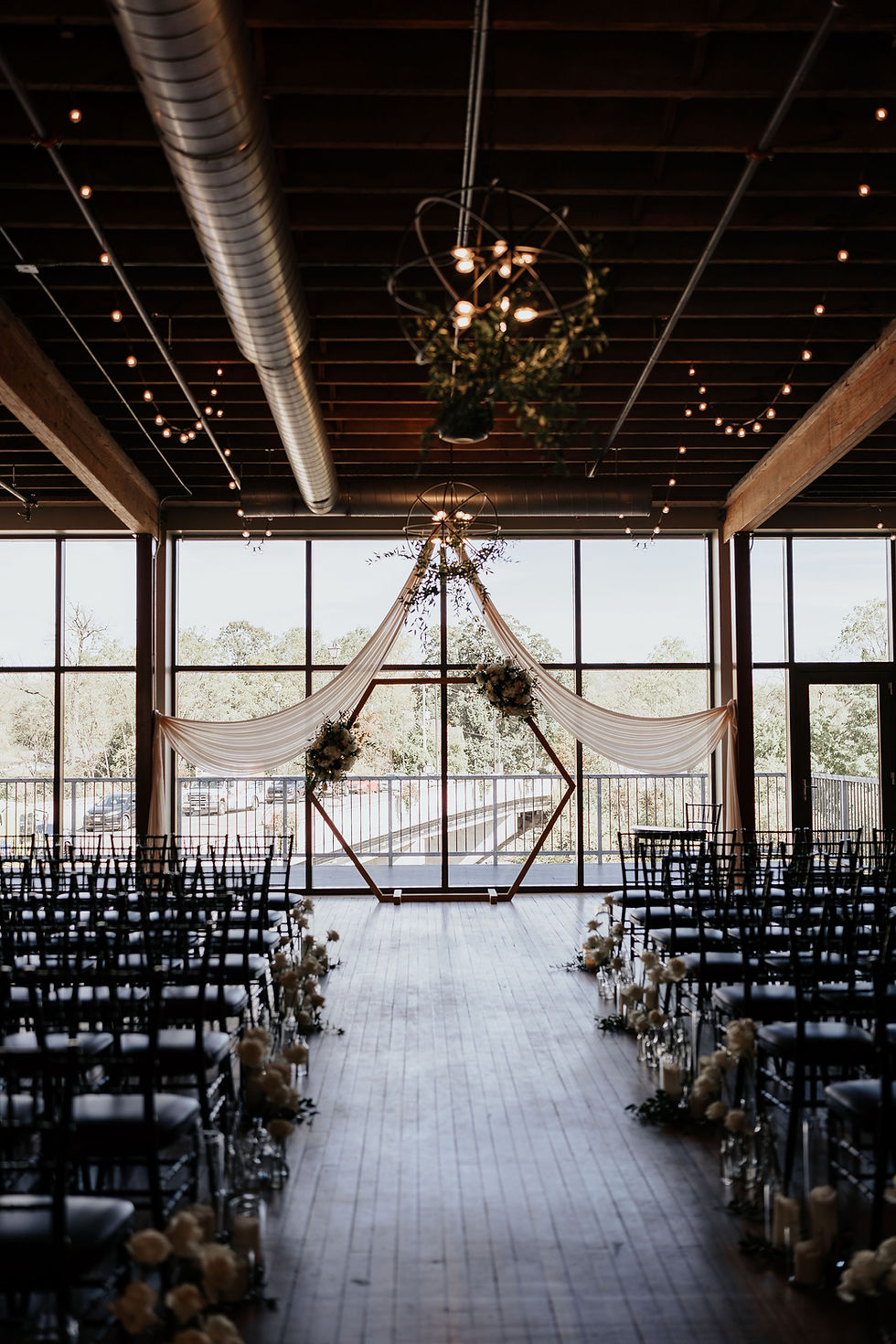 Wedding venue with wooden hexagonal arch draped in white fabric, floral decor, and empty chairs. Soft lighting and large windows in background. bride and groom portraits River’s Edge at The Arlington Wedding – Paris, Ontario