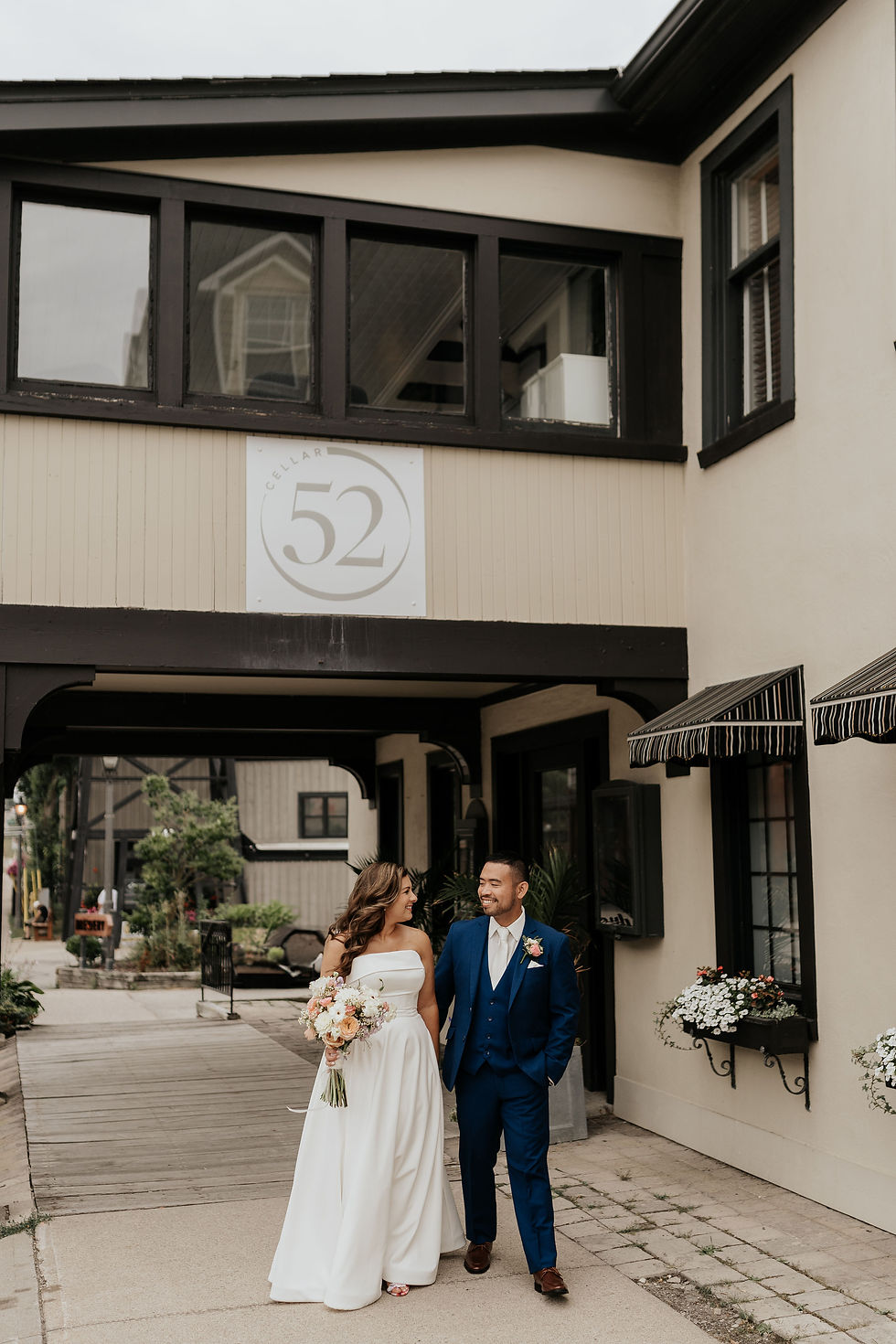 Bride in white dress and groom in blue suit smile walking together outside Cellar 52. Surrounded by flowers and black-trimmed windows.