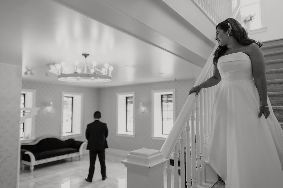 Bride in a white gown on stairs, looking at a groom in a suit in a bright room with elegant chandelier and windows. Romantic mood. cellar 52
