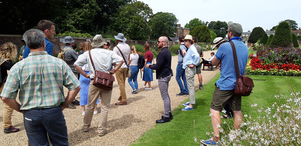 Tour group in a formal garden stands on a gravel path, many wearing hats and backpacks, listening and looking around under sunny skies.