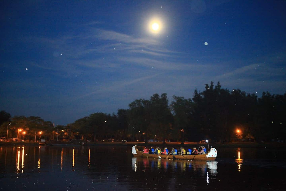 A group of individuals canoeing at night time in the summer. The moon is located above the group.