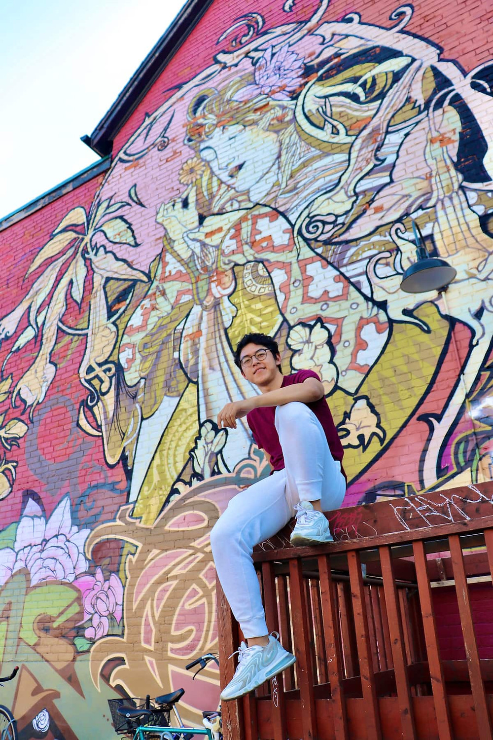 An individual posing in one of the photography spots found in Kensington Market's Photography Walk, located in the heart of downtown Toronto. There is a painting of a person and abstract shapes on a brick building in the background.