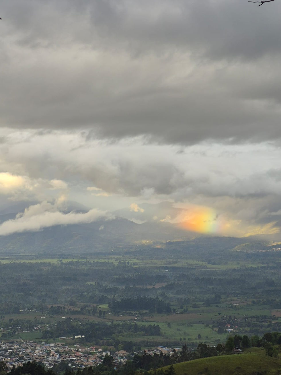 iew over the Sibundoy Valley, with a town and green fields below cloud-covered mountains and a warm glow on the horizon.