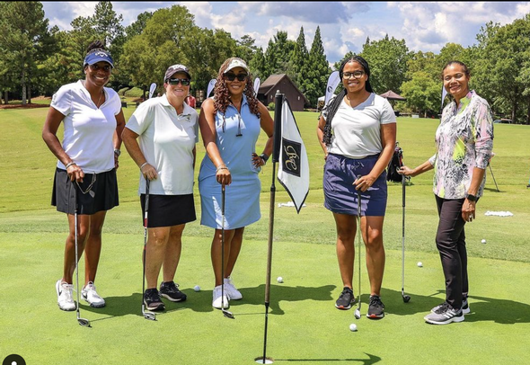 Five smiling women posing with golf clubs on a green.