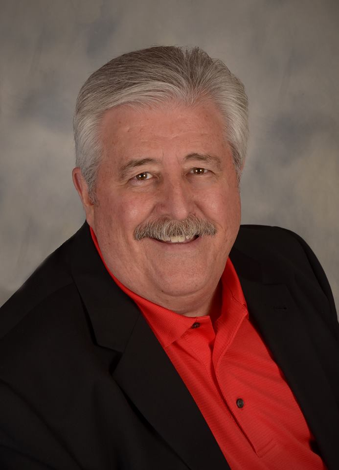 Smiling man with grey hair, mustache, red shirt, and black blazer.