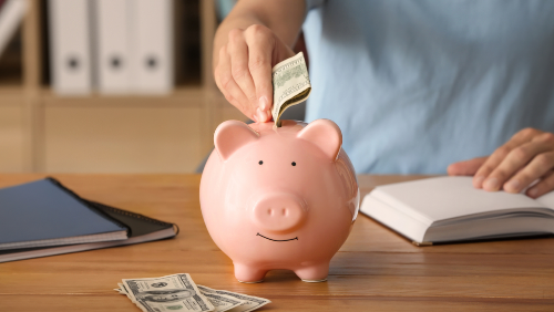 Person putting a folded dollar bill into a pink piggy bank