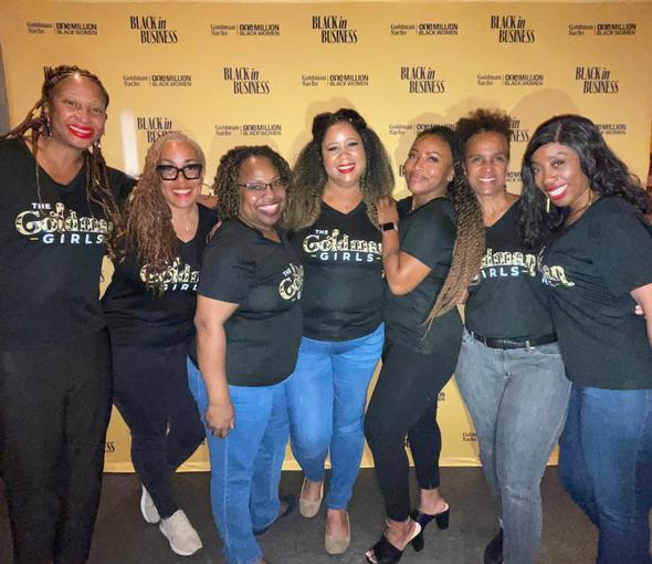 Seven women in matching black t-shirts smiling at an event