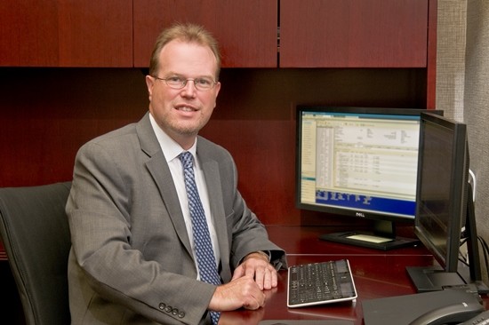 Man in a grey suit sitting at a desk with two monitors.
