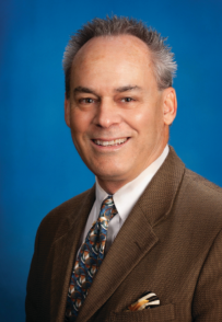 Smiling man in a brown suit and patterned tie.