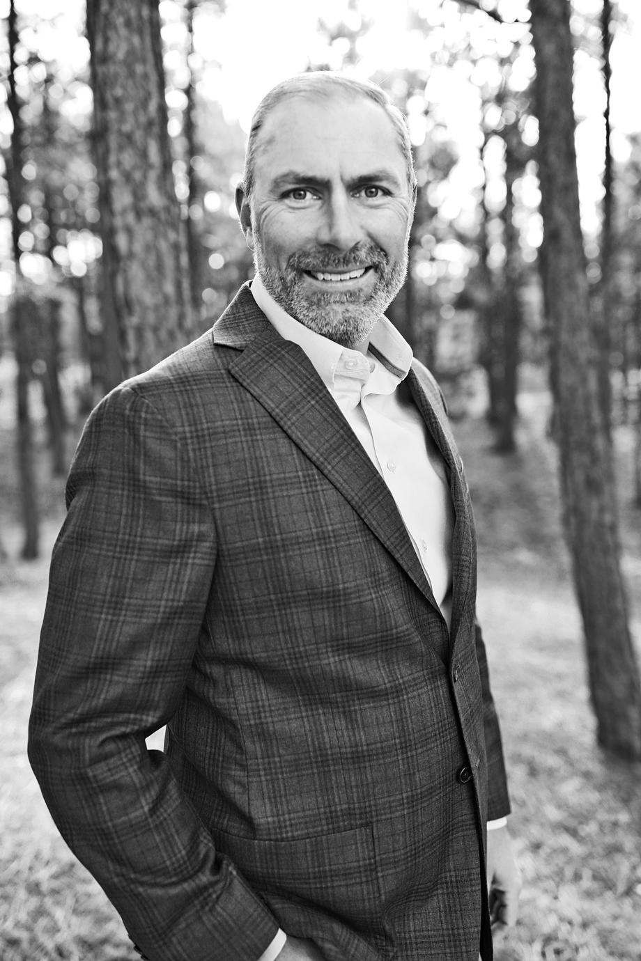 Man in a plaid suit jacket smiling in a forest setting.