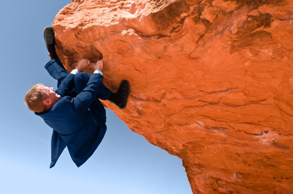 Businessman in a suit hanging from a steep rock cliff.