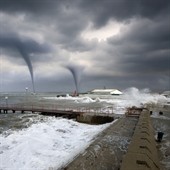 Two water spouts forming over stormy seas near a pier.