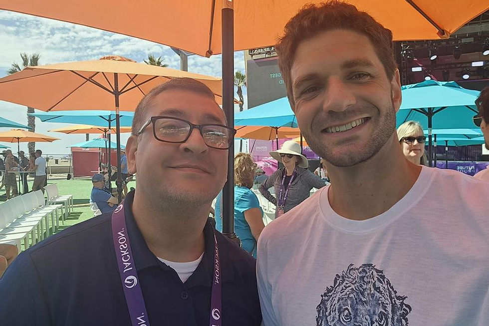 Two men smiling at an outdoor event under umbrellas.