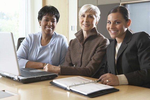 Three professional women smiling around a laptop and planner.
