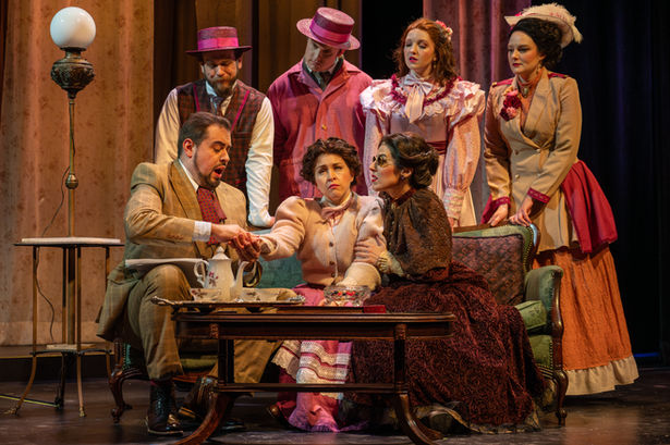 Three performers playing John Macy, Helen Keller and Anne Sullivan sit on the sofa as John fingerspells his name into Helen's hand. Her choir stands around them commenting on the character qualities of John Macy that are lacking.