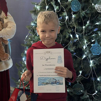 Nagrodzony chłopiec prezentuje otrzymany dyplom, stojąc na tle choinki.