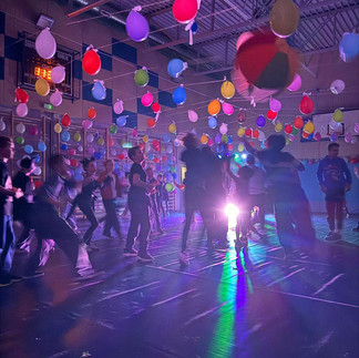 Dzieci w strojach karnawałowych tańczące na sali udekorowanej balonami, w kolorowym świetle dyskotekowych reflektorów.